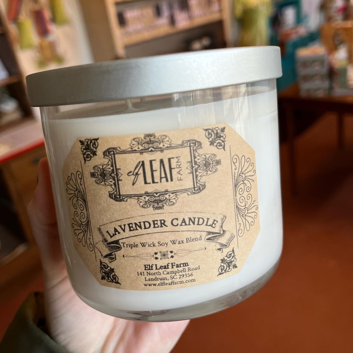 A white candle with the label "Elf Leaf Farm Lavender Candle" is held close to the camera. 