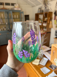 A stemless wine glass with a handpainted lavender motif is held in a person's hand. 